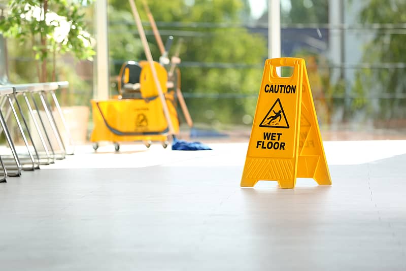 a wet floor sign sits on a gray floor with a mop bucket from a cleaning company in the background 
